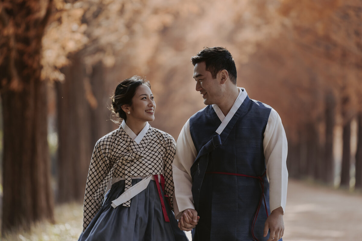 the couple holding hands and staring at each other while wearing their hanbok in a pre-wedding shoot in metasequoia road in damyang south korea