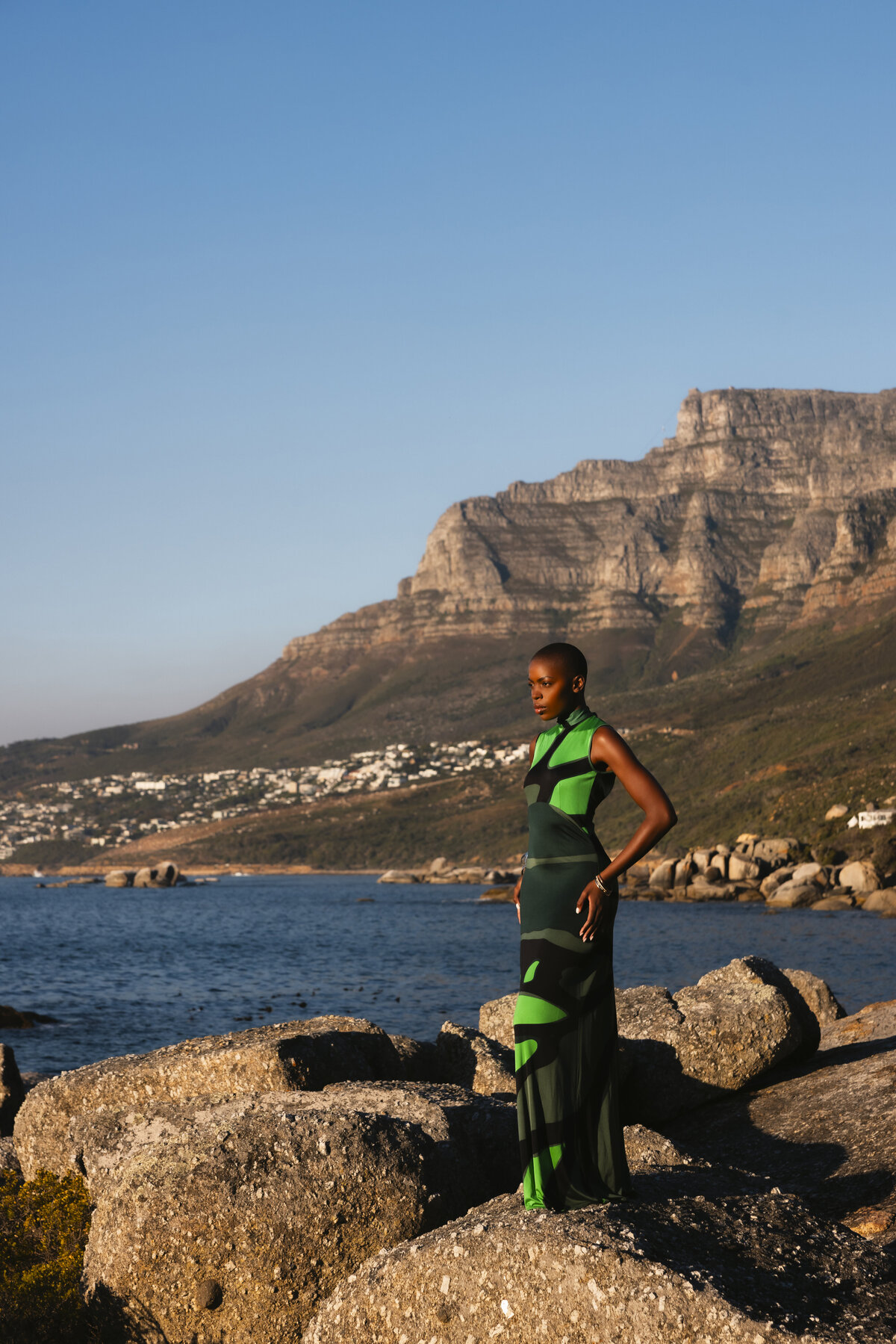 Model in a bold green and black dress standing confidently on seaside rocks, with the dramatic cliffs of Cape Town’s Twelve Apostles mountain range in the background under clear blue skies.