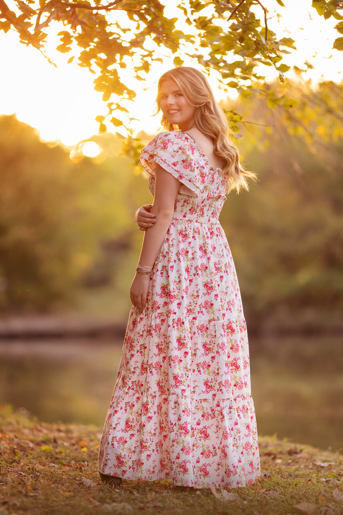 A young woman in a floral dress smiles over her shoulder as the golden sunset light highlights her hair and the soft background.