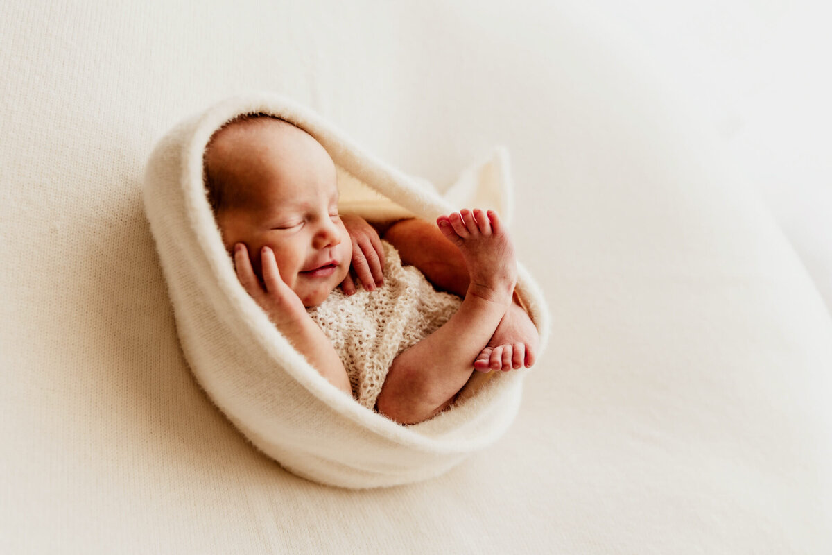 Baby who is wrapped in a white swaddle smiles while laying on a cream blanket and swaddled, OKC newborn photos.