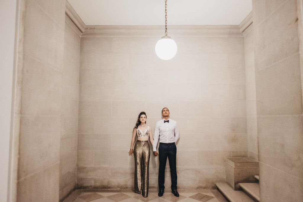 The photo was taken at Washington D.C., specifically The Mellon Auditorium, known for being one of the best luxury DC venues with European flair by photographer Sarah Bradshaw. In the image, a couple stands holding hands in a plain interior with beige walls and a tiled floor. The woman is dressed in a glittery gold outfit, creating a striking contrast against the neutral background, while the man exudes a classic charm in a white shirt, dark pants, and a stylish bow tie. A single round light elegantly hangs above them, illuminating their connection in the setting.