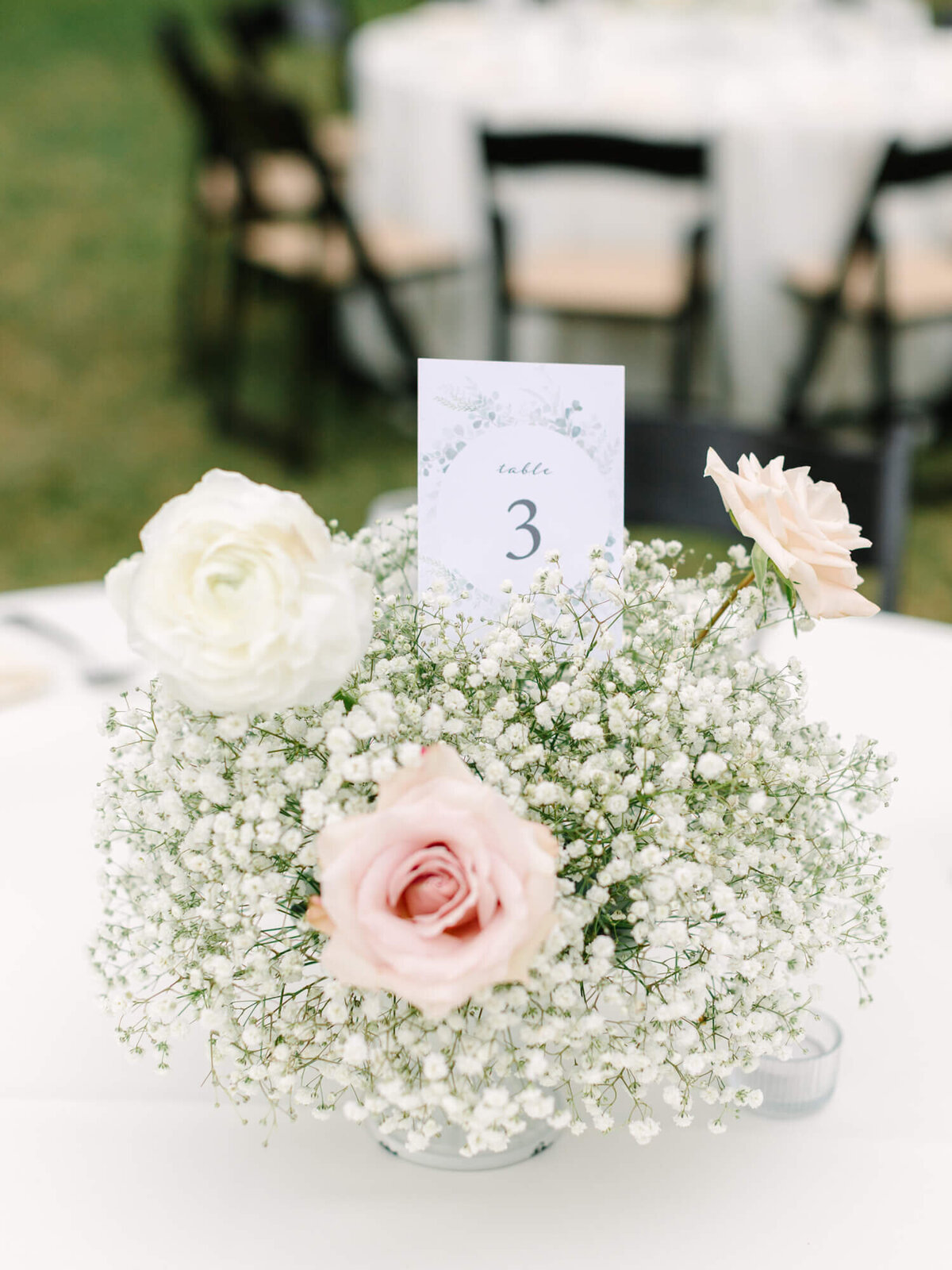 A centerpiece with pink and white roses and baby's breath surrounds a "Table 3" card on a round table at an outdoor event. Soft, elegant atmosphere.