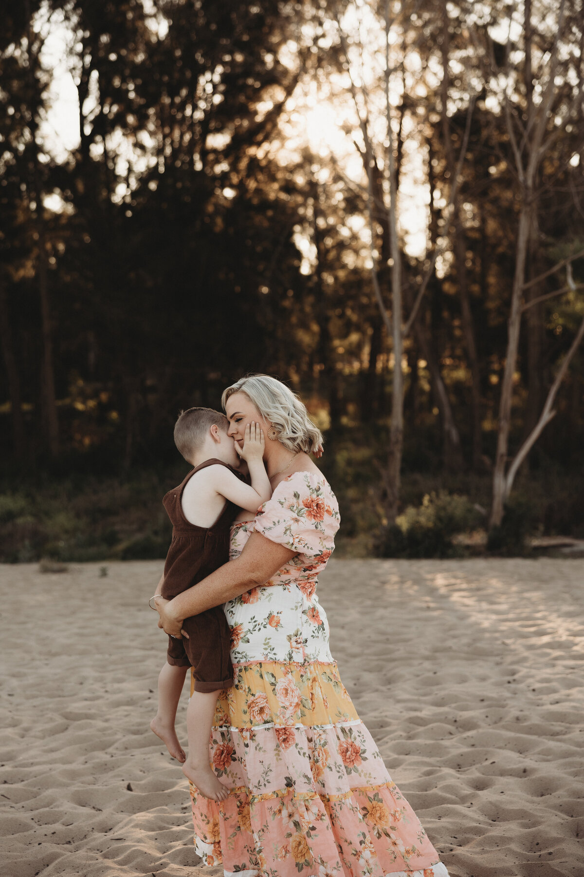 A woman in a floral dress holds a young child while standing on a sandy area with trees in the background, beautifully captured by an Illawarra newborn photographer.