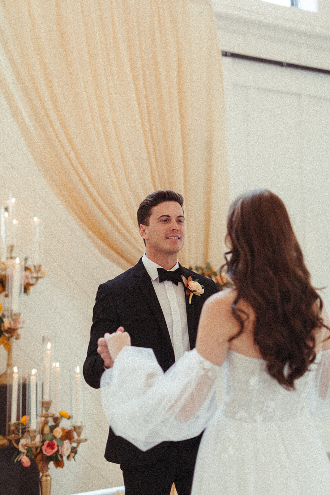 Bride and groom wearing a black tuxedo and white wedding gown dance in a room filled with flowers arrangements and candles