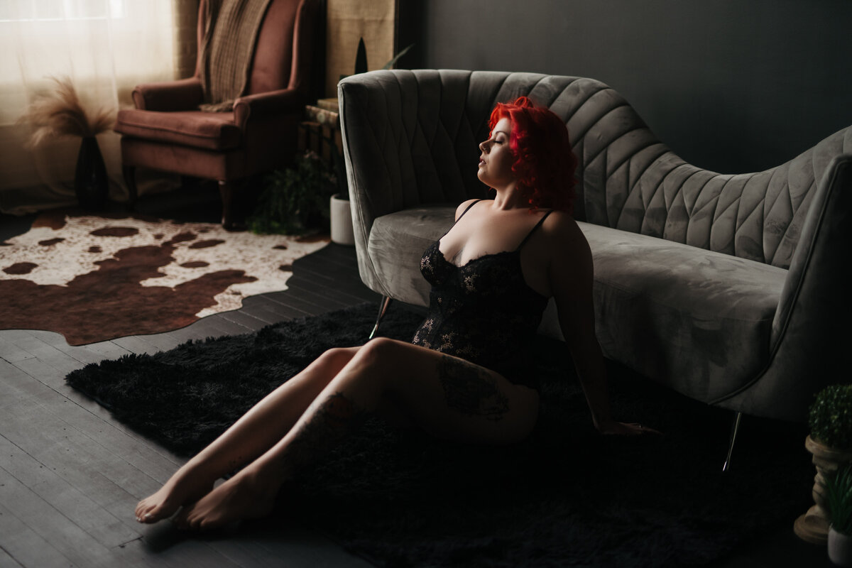 A woman with red hair sits on a floor leaning against a grey couch on a black rug posed by a New England Boudoir Photographer
