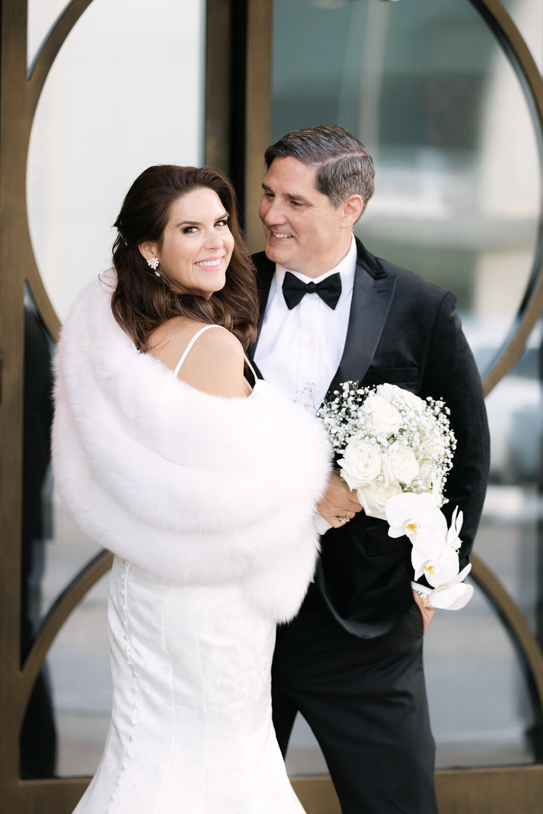 Winter wedding portrait in downtown Dallas with the bride in a white fur cover and sleek gown, admired by the groom