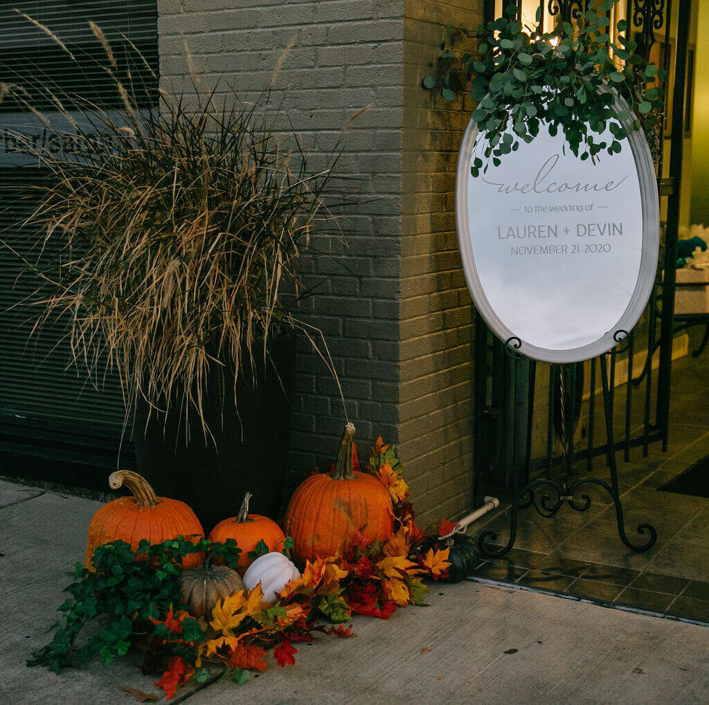 A wedding entrance with a welcome sign for Lauren and Devin. The scene is warmly lit, decorated with greenery and autumn wreaths.