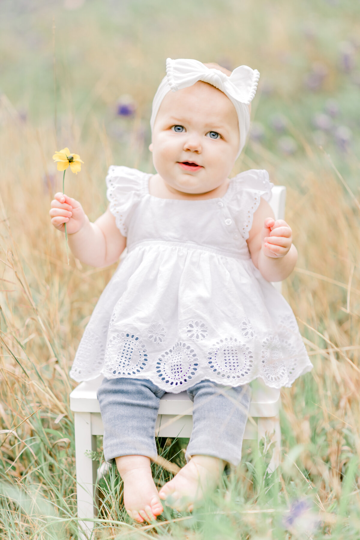 Baby girl sitting on a chair in Texas wildflowers holding a flower and looking at camera