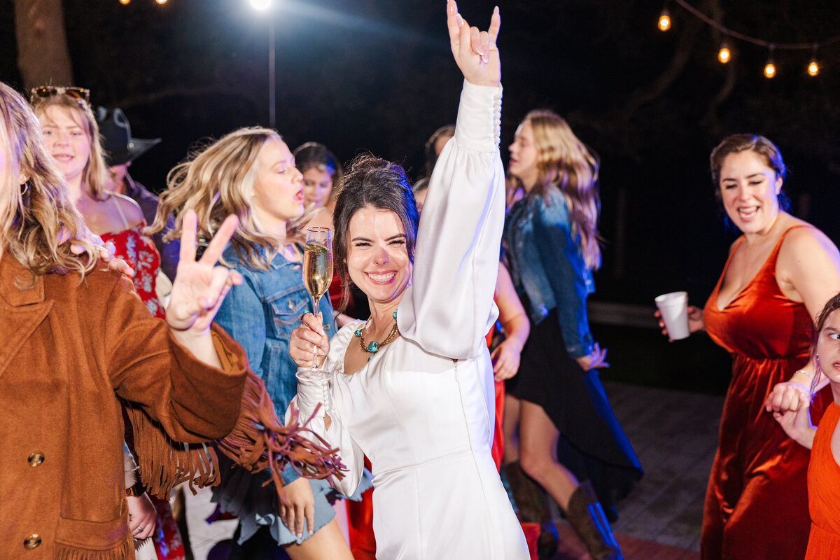 San Antonio wedding photographer captures the bride at the reception all excited and dancing. The bride is looking at the camera with a hand in the air in a rock and roll sign and a drink in hand.