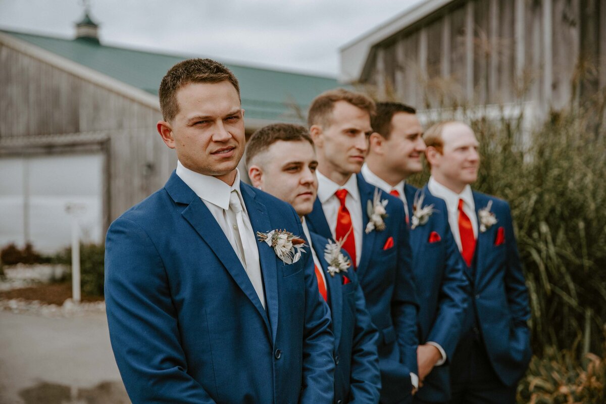 Groom and his groomsmen standing on an Exeter, Ontario farm with a rustic barn behind them. He is waiting for his bride to come down the aisle. The groom is smiling at the camera and the groomsmen are looking off in the distance.