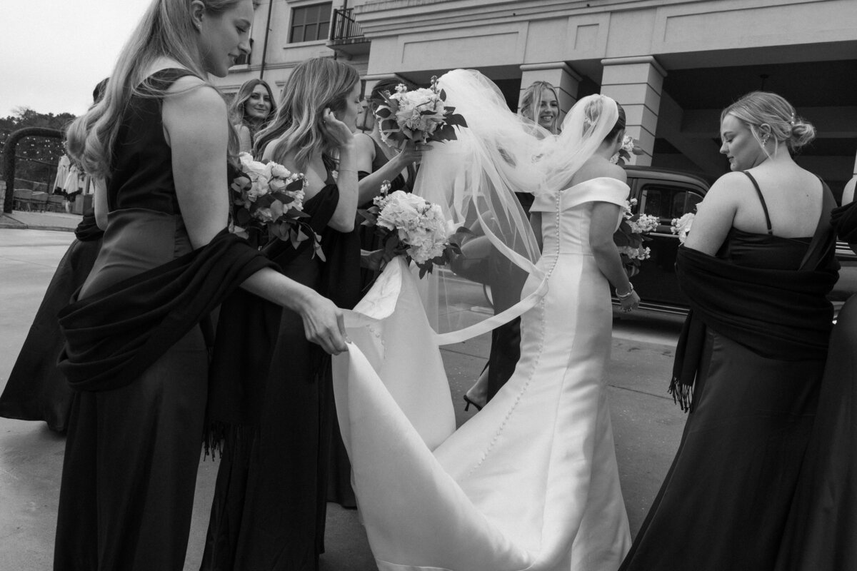 A bride walking away while all her bridesmaids help her by holding her veil and dress.