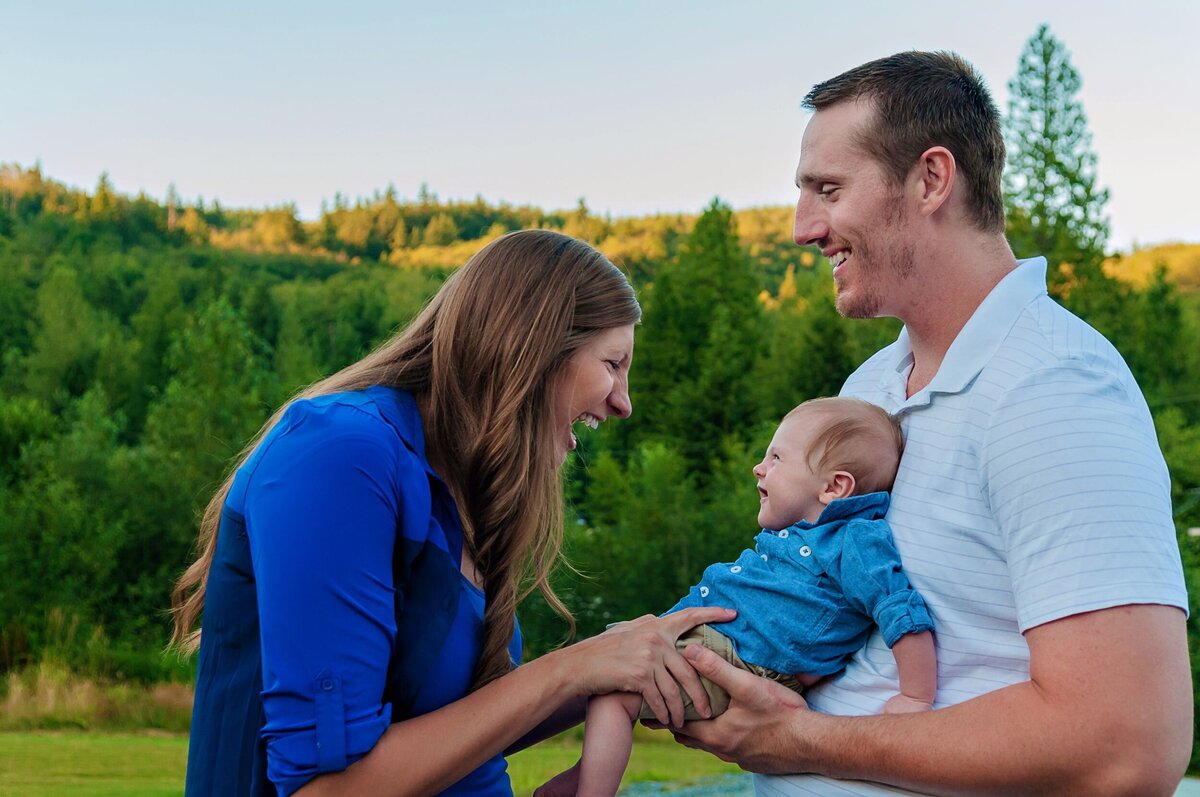 Dad holding baby and mom laughing with the baby, all dressed for summer outdoors with beautiful green trees in the background