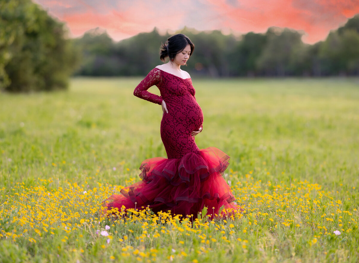 Elegant maternity photo of a woman in a red lace gown standing in a field of yellow wildflowers in Leander, Texas, with a vibrant sunset sky.