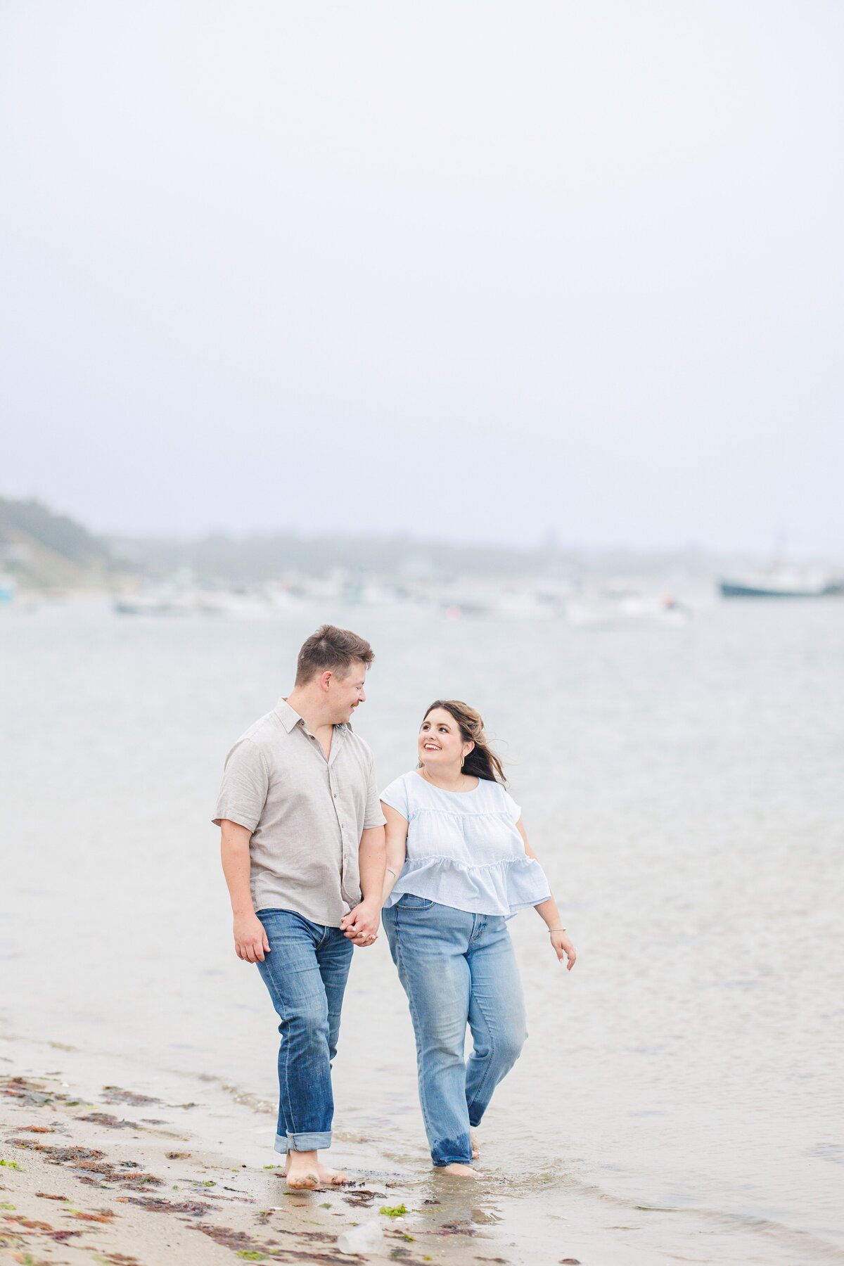 Couple walking hand in hand along the shoreline, smiling at each other, with gentle waves lapping at their feet and boats in the background at Chatham Bars Inn in Chatham, Massachusetts — representing the portfolio of a Cape Cod engagement photographer.