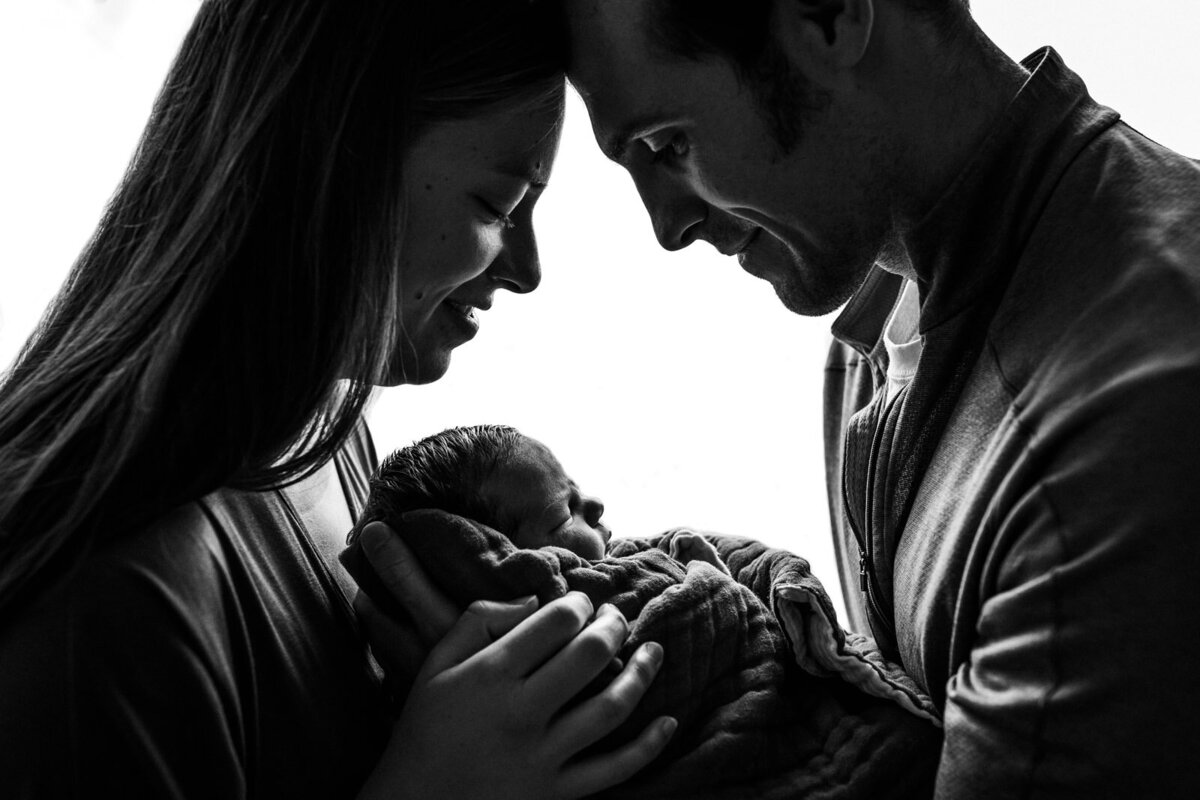 mom and dad touching foreheads and looking down at newborn they're holding together and smiling