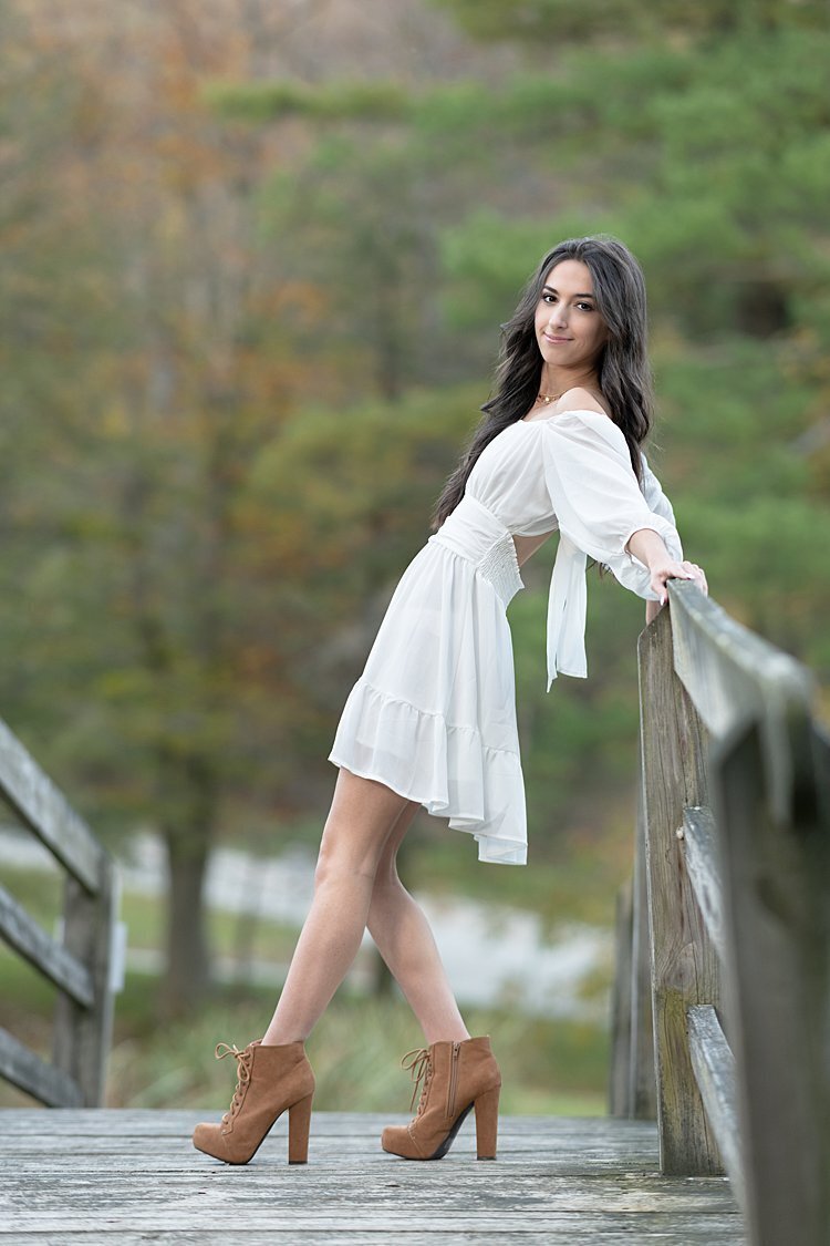 High school senior girl in white dress leaning against wooden railing on bridge