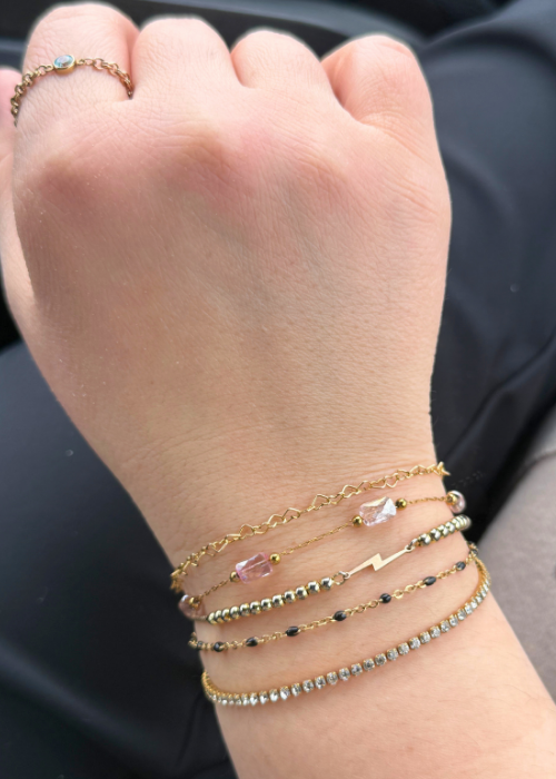 A wrist stacked with multiple gold and gemstone bracelets, featuring beads, lightning bolt, and crystal details, showcasing a permanent jewelry style.