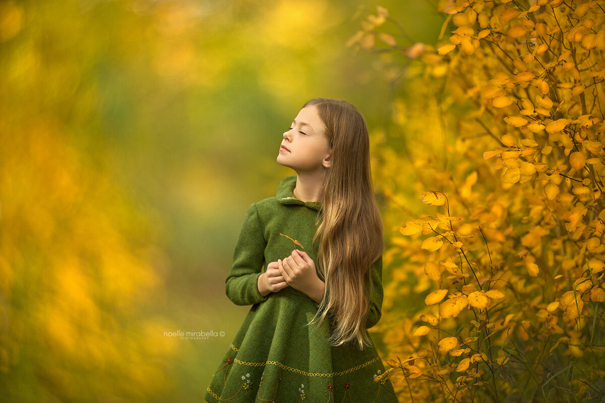 Girl in green wool dress standing next to a golden autumn tree.