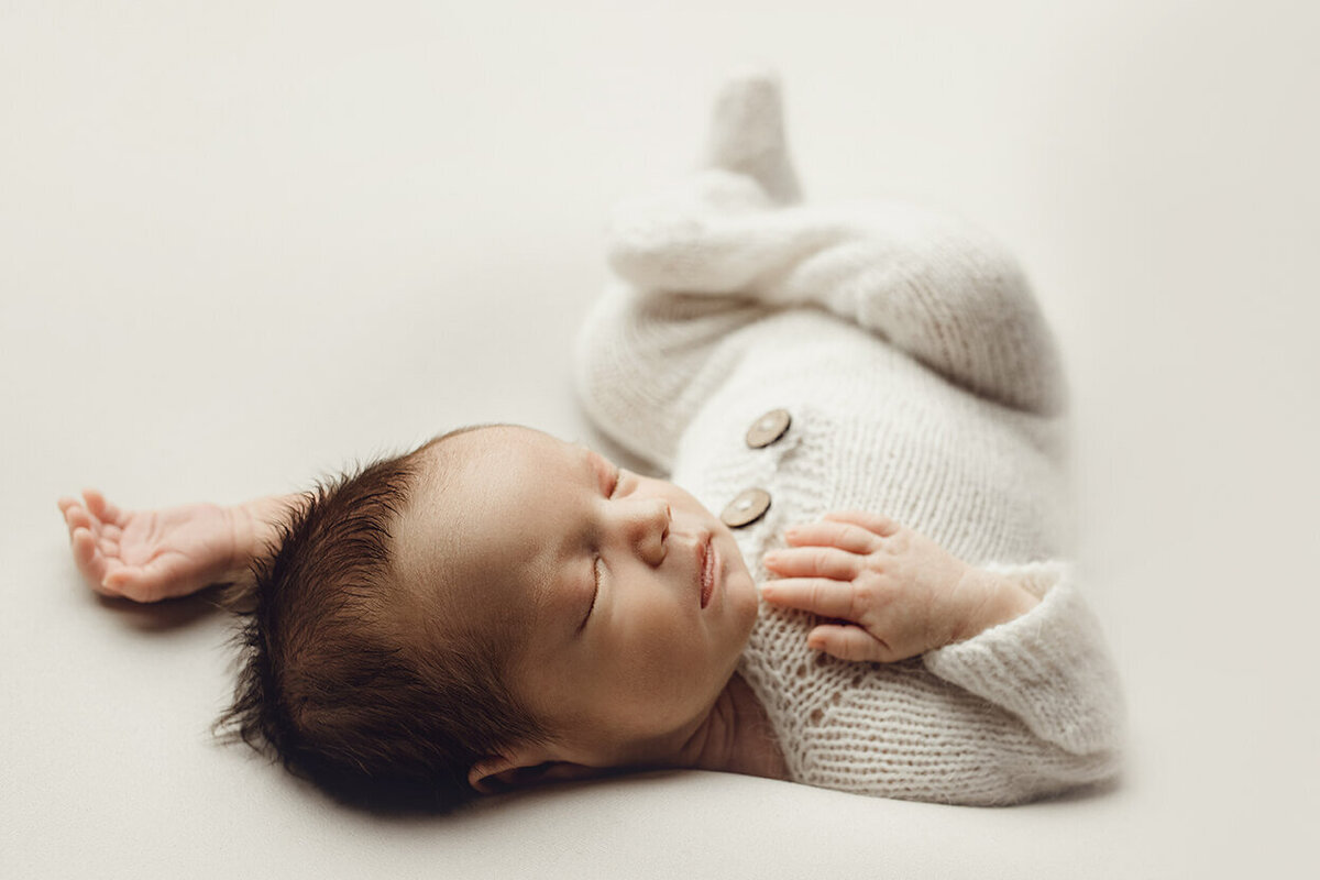 very relaxed pose of a baby boy sleeping on his back in a white knitted romper