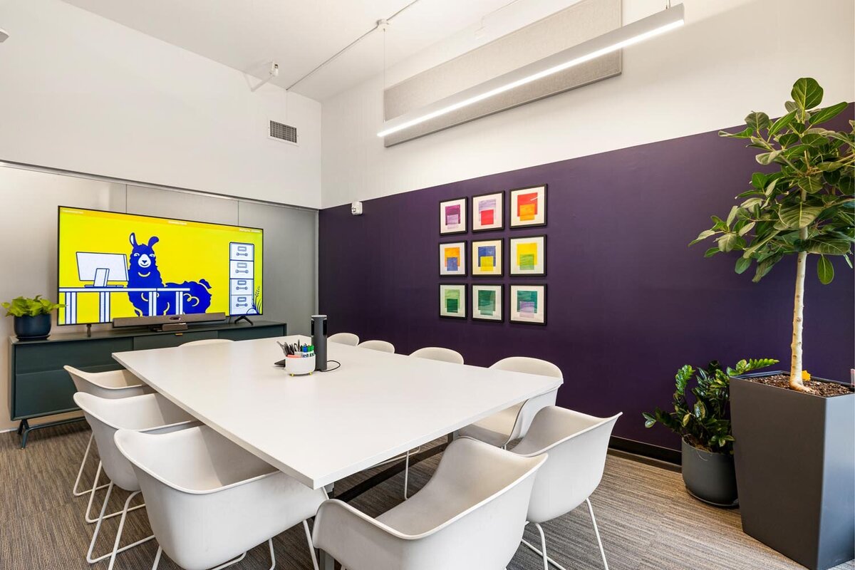 Conference room with white meeting table, purple feature wall, colorful framed art, and TV displaying mascot.
