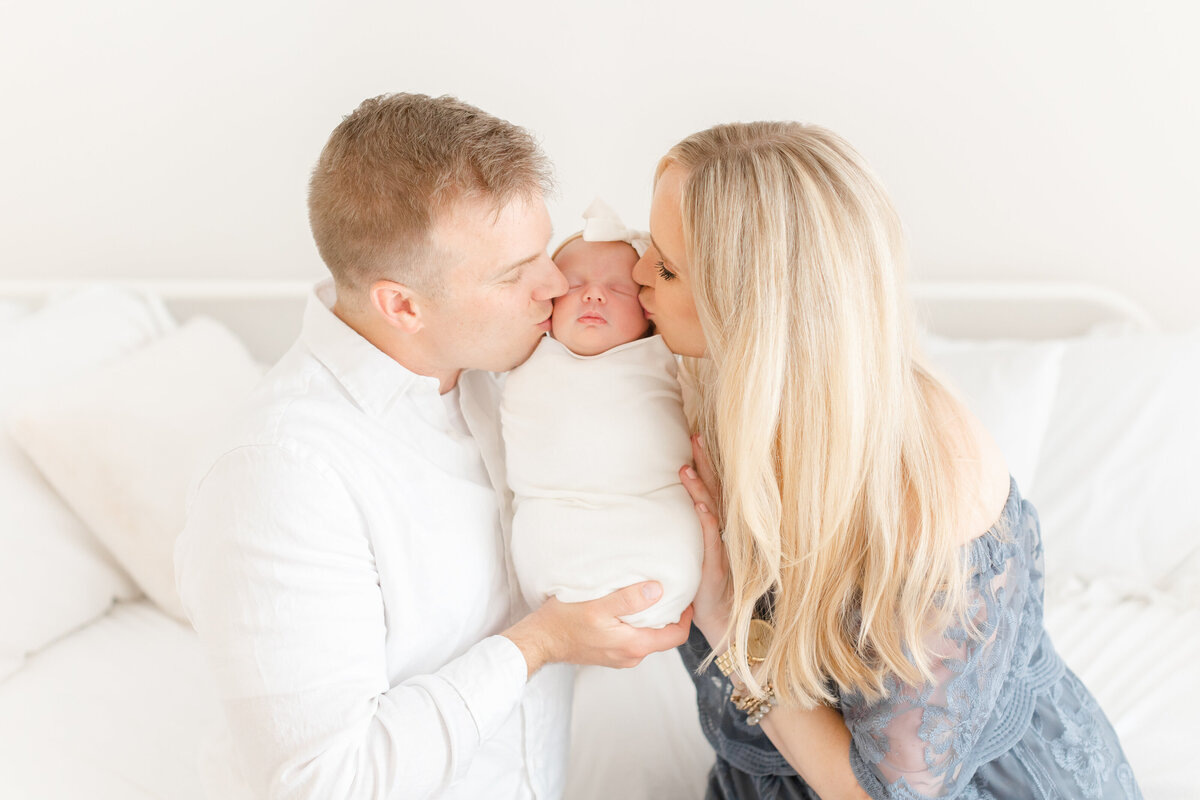 A blonde mother and brunette father kissing their baby in their arms on a white bed by Washington DC Maternity photographer