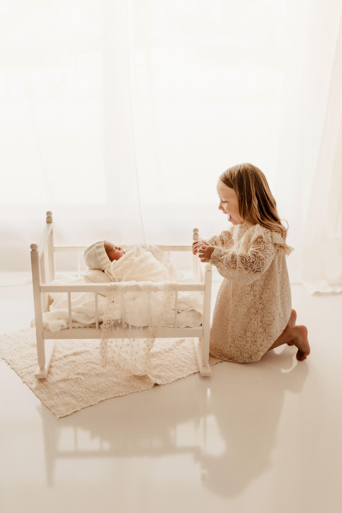 Girl kneeling down on a white reflective floor with a window behind her smiling down at her baby sister.