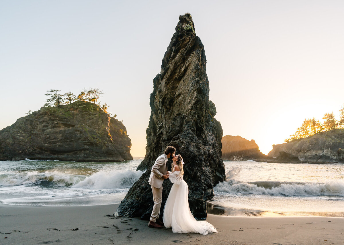 A couple in their wedding attire face each other perfectly centered by their adventure elopement photographers in front of a jagged sea stack. Crashing blue ocean waves roll behind them as they embrace in the golden light of the sunset on this rocky coast line. 