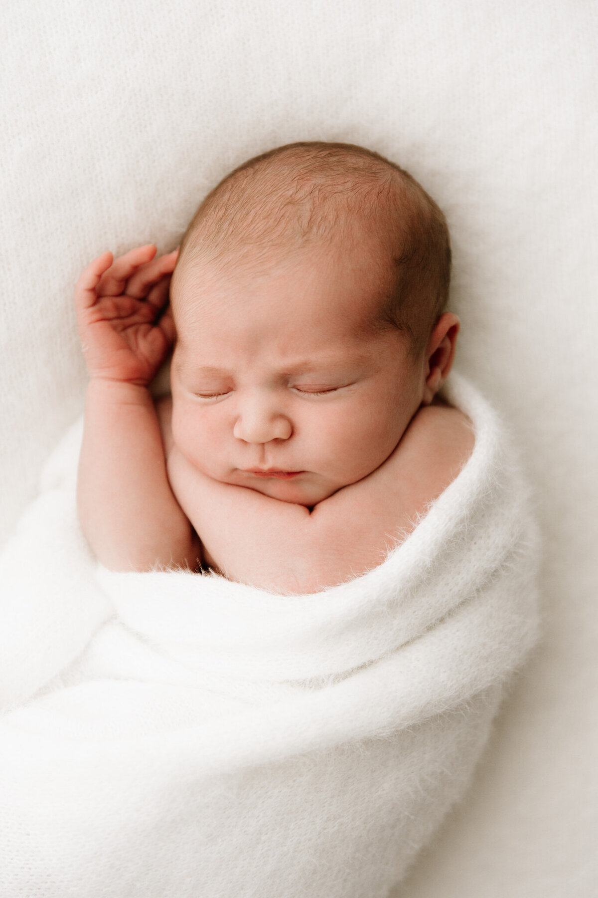 A close-up portrait of a newborn baby peacefully sleeping with arms gently tucked under the chin, wrapped in a soft white blanket on a textured white surface. The baby's delicate skin and fine hair are softly illuminated by diffused studio lighting. Photographed in a Sydney newborn photography studio using artificial light.