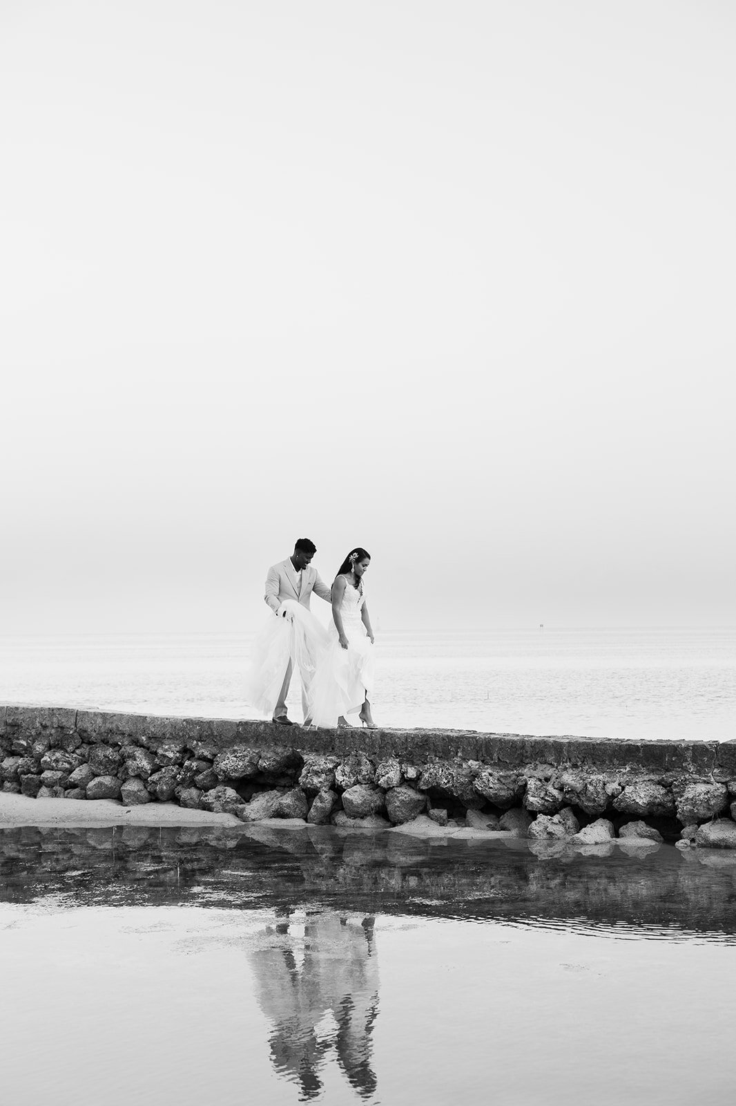 Andressa and Jonathan walk hand in hand along a rocky shoreline, captured in a timeless black and white image at their Islamorada wedding. Photography by Claudia Amalia Wedding Photography.