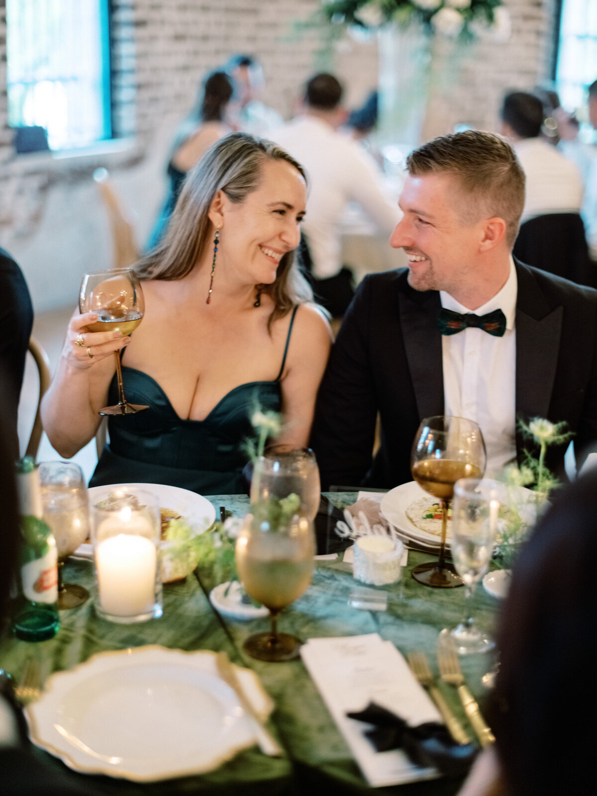 A stylish couple shares a moment at an elegant dinner event, surrounded by beautifully set tables, soft lighting, and a festive atmosphere that elevates their connection.