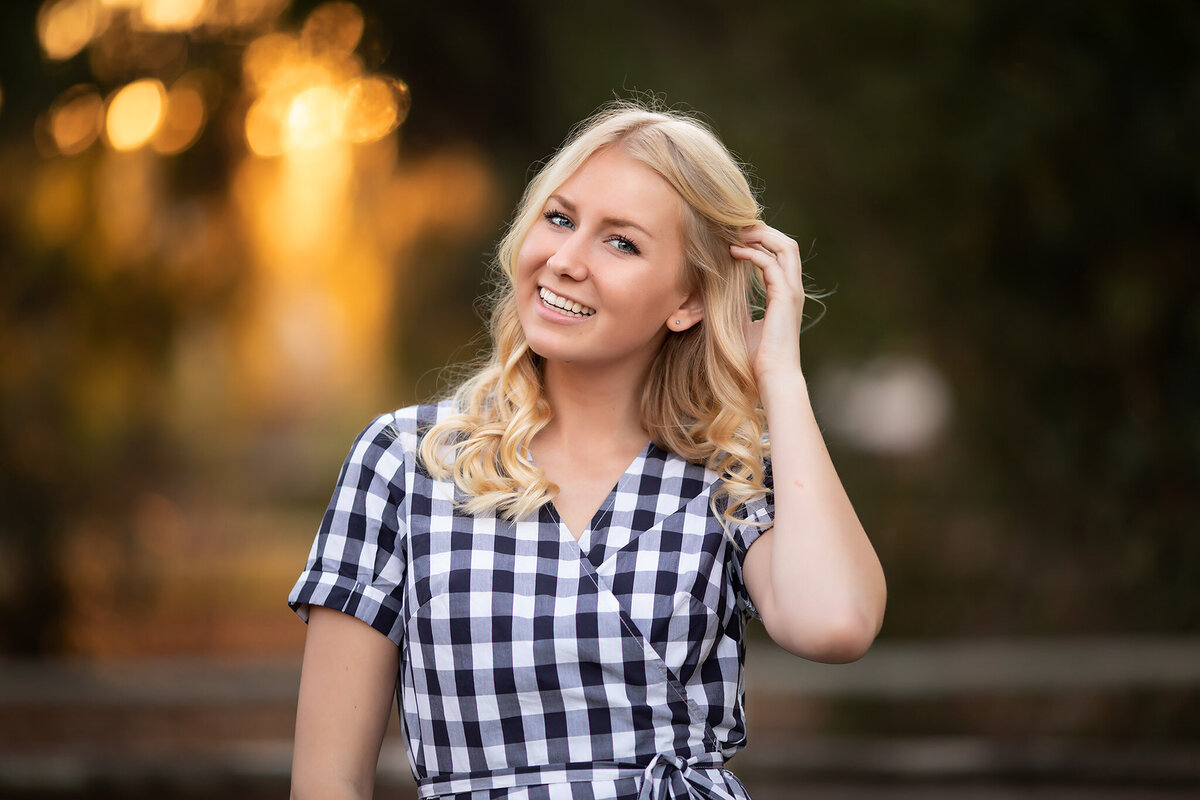 A young woman with blonde curls smiles in a checkered dress, captured by a Dallas photographer with a glowing sunset backdrop.