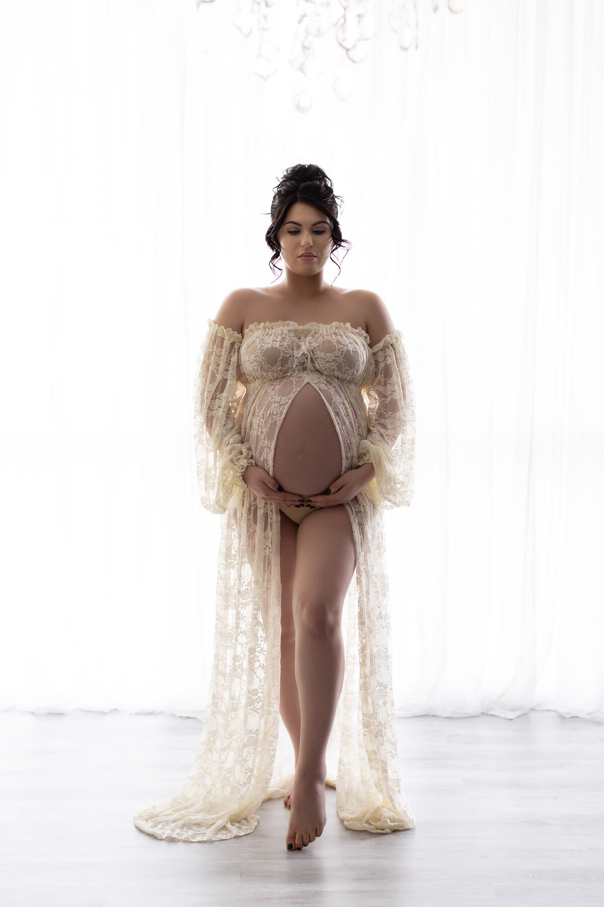 A serene maternity photograph featuring a woman standing gracefully in front of soft white curtains, wearing a luxurious lace gown with off-the-shoulder, flowing sleeves and intricate detailing. The gentle backlighting highlights her silhouette, evoking a dreamy and ethereal quality. Captured by Lindsay Walden Photography.