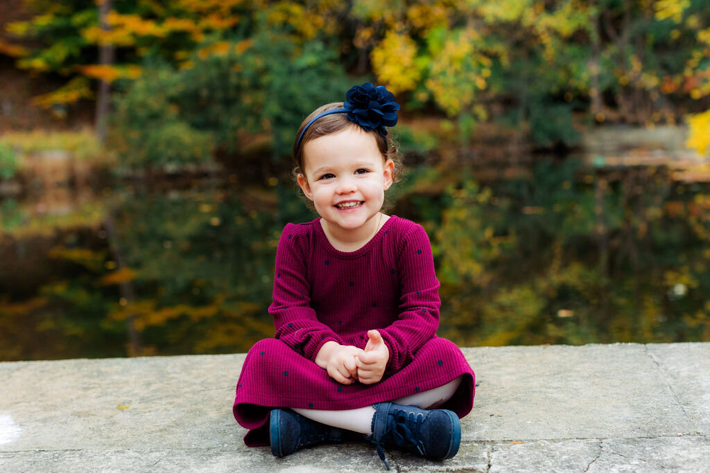 A child sits and smiles outdoors in a raspberry-colored dress.