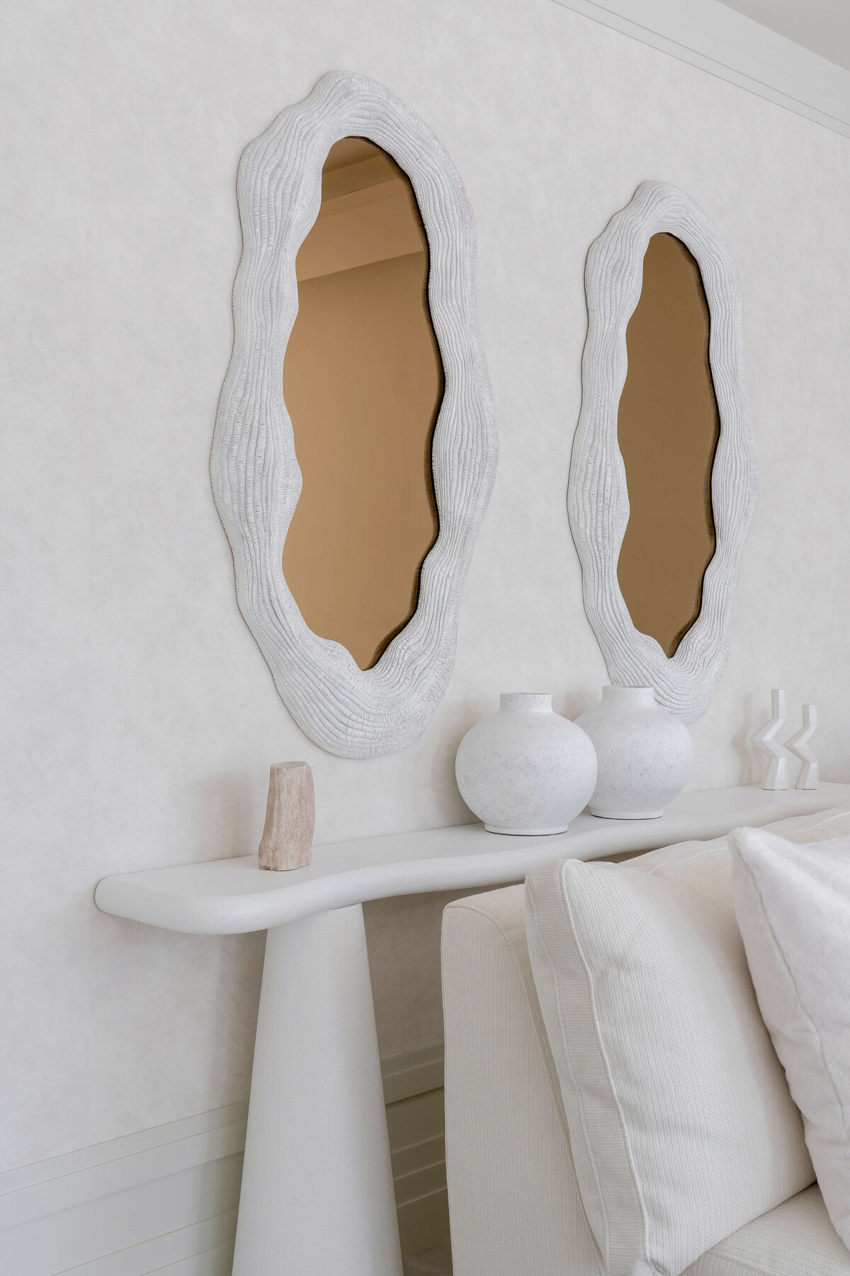 Behind a white couch is a white console table adorned with neutral colored decor. Above the table are two large, contemporary mirrors.