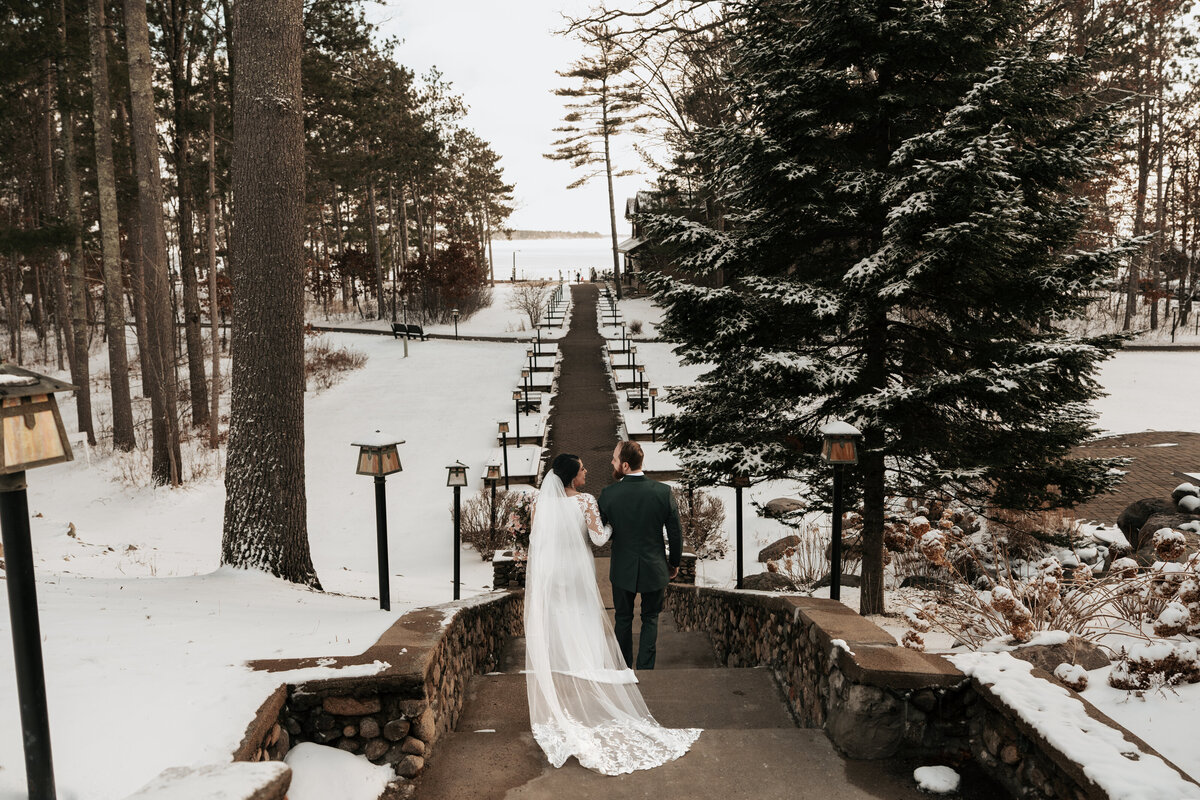 Newly married couple stands on a snow-covered stone path overlooking a winter lake, surrounded by evergreen trees in a scenic Minnesota elopement setting. 