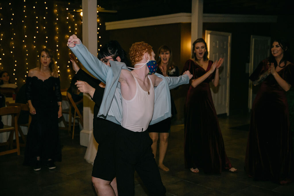 A group of people in formal attire dance. In the center, a person with red hair in a tank top and open jacket dances energetically, wearing a colorful face mask. Twinkling string lights are in the background.