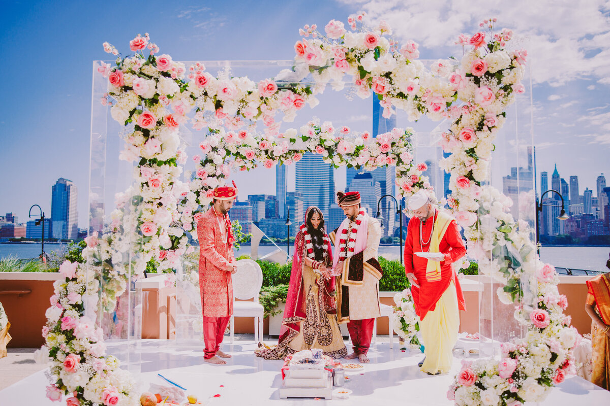 Indian bride and groom at  the wedding during their hindu ceremony under a floral see-through mandap with new york city skyline at the jersey city hyatt