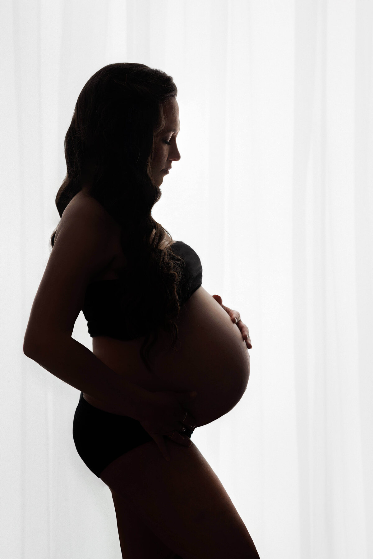 pregnant mother wearing a black bandeu top with her pregnant stomch showing in front o a white  backlit curtain at her maternity session in northern va