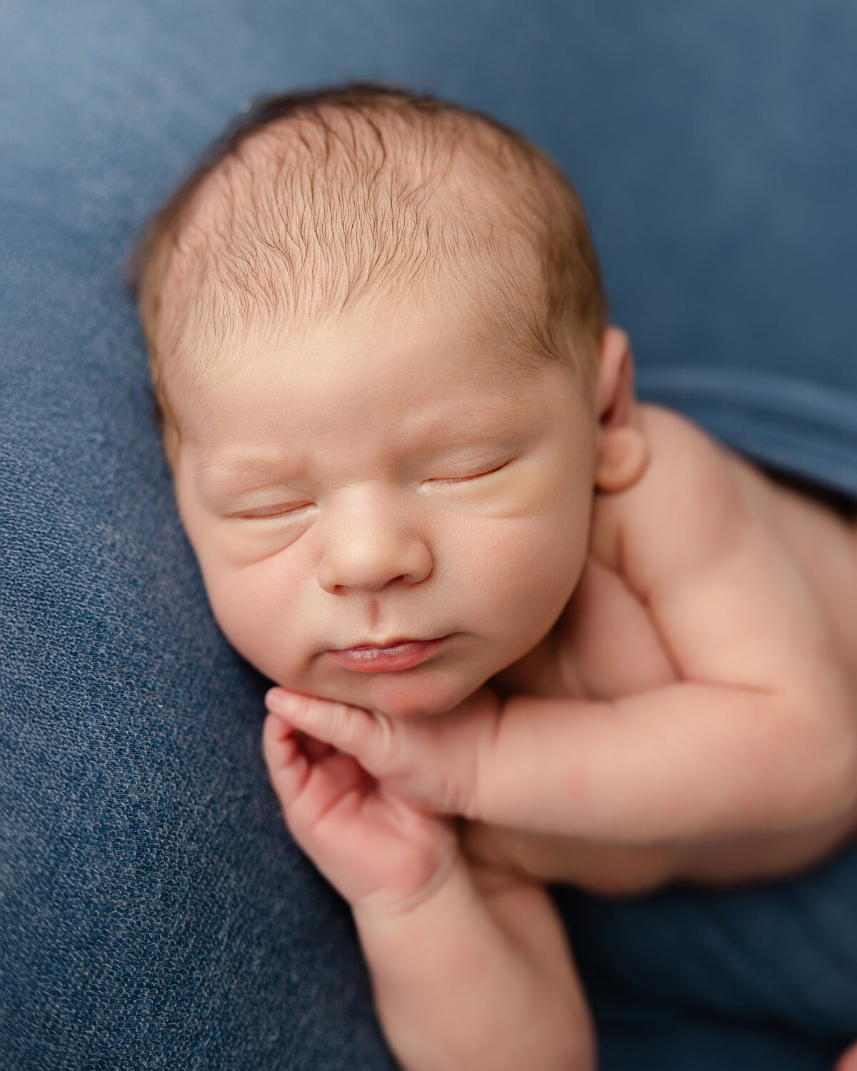 Newborn baby boy sleeping peacefully on a soft blue blanket with his hands gently tucked under his chin. Captured by Inland Empire newborn photographer Kaitlyn Dawn Photography.