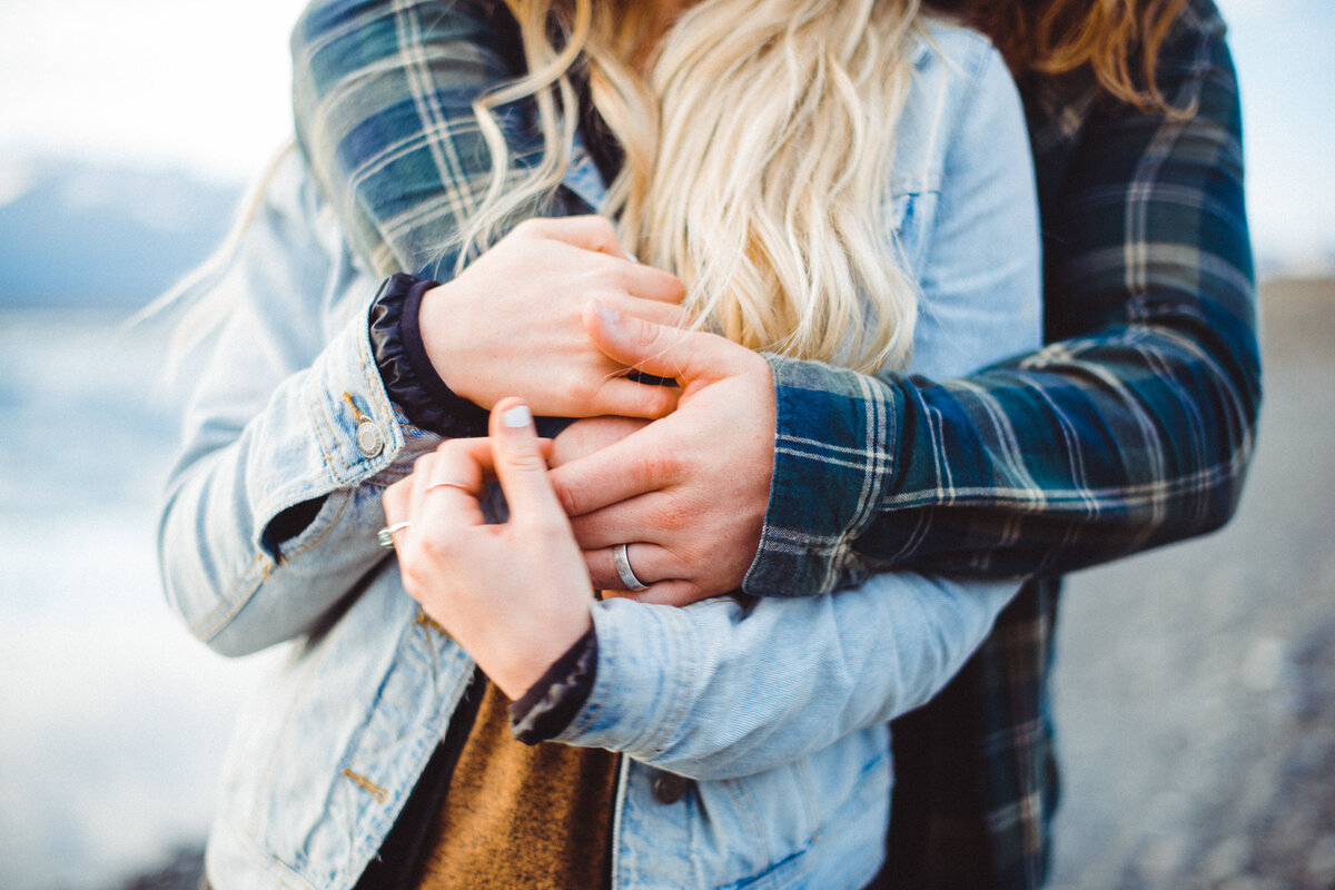 Intimate elopement ring detail in Iceland. Close-up of couple's intertwined hands wearing wedding bands, dressed in cozy layers with denim jacket and plaid flannel against misty Icelandic landscape. Adventure elopement photography capturing authentic moments in the Nordic wilderness. Destination wedding details with moody, romantic aesthetic.