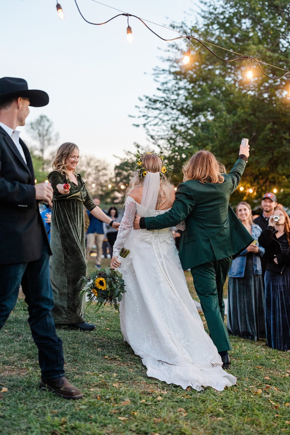 Shot of a bride and groom from behind as they are announced to enter their wedding reception in Fort Worth, Texas. The bride is on the left and is wearing a long, flowing, intricate, long sleeve, white dress with a veil and flower crown while holding a large bouquet. The groom is on the right and is wearing a green suit. Guests celebrate and welcome them in on all sides.