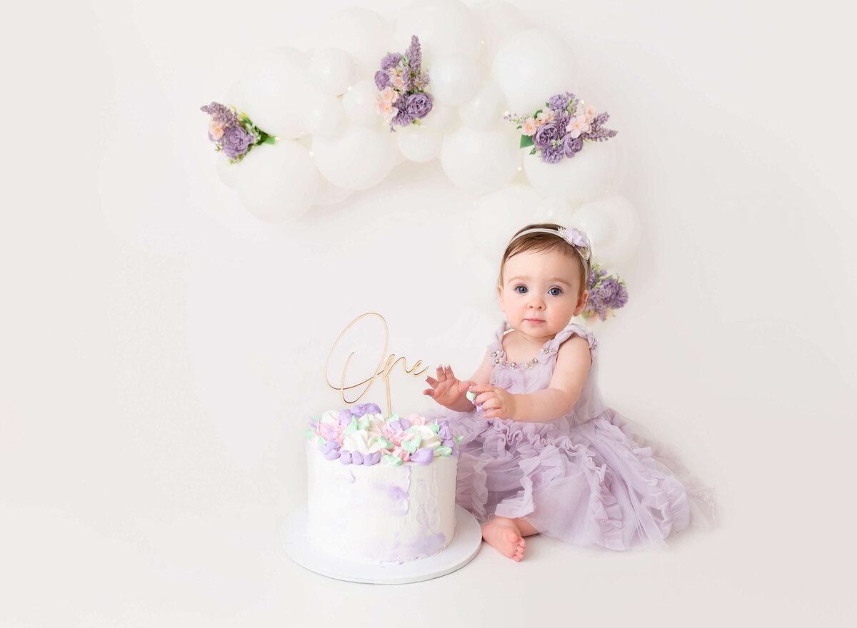 Baby girl celebrating her milestone with a cake smash session, surrounded by white balloons and lilac flowers, captured in a NYC cake smash photography session for timeless memories.