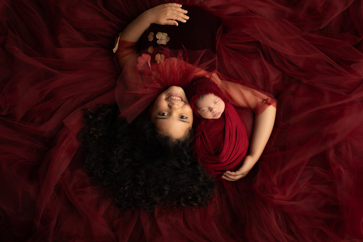 Older sister smiles while laying next to swaddled newborn baby in deep red tulle fabric, captured by Dallas newborn photographer.