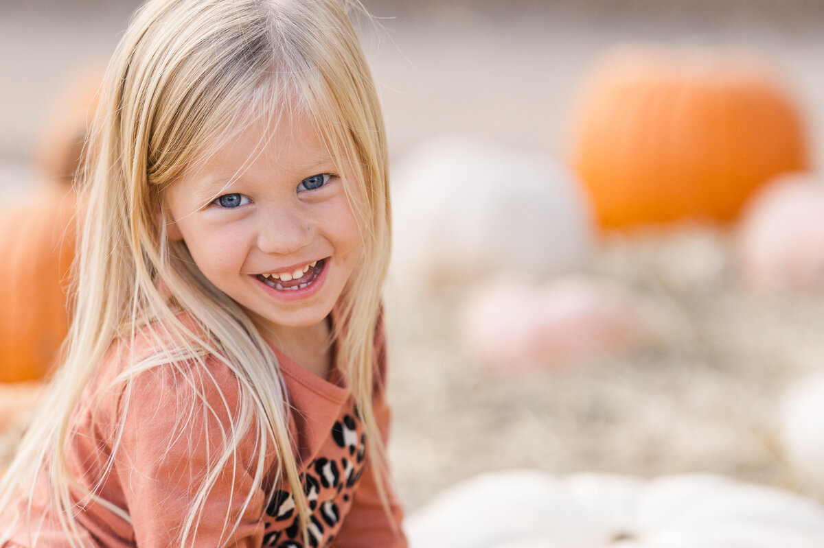 3-year-old-gilr-smiles-at-the-camers-while-sitting-on-a-pumpkin-at-the-pumpkin-patch