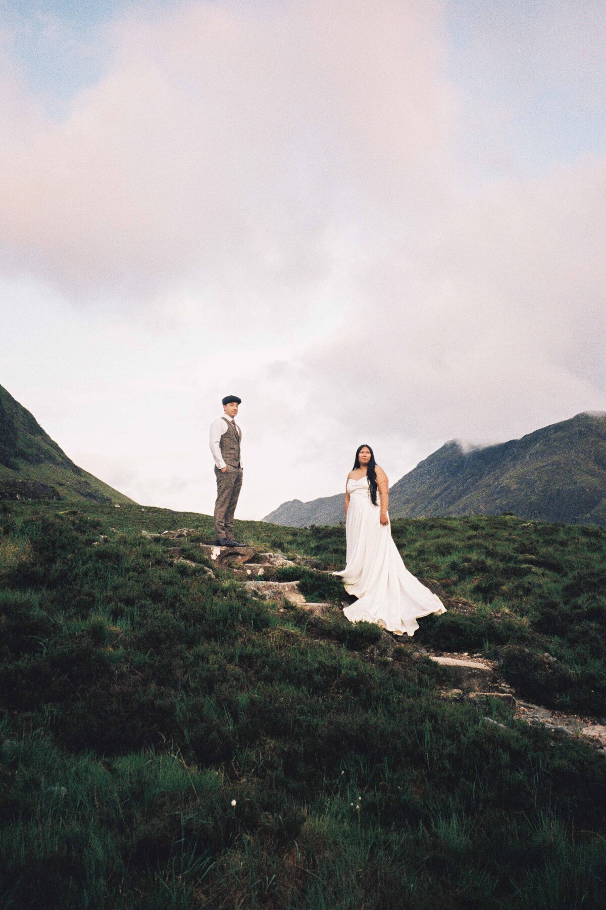 A bride and groom are photographed standing around 4 meters apart. The 35mm film photograph shows them standing on a stone staircase in the middle of the Glencoe landscape. They wear wedding attire.