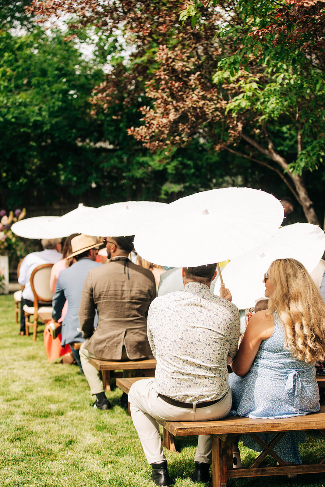 People seated on benches holding white umbrellas at an outdoor wedding ceremony.