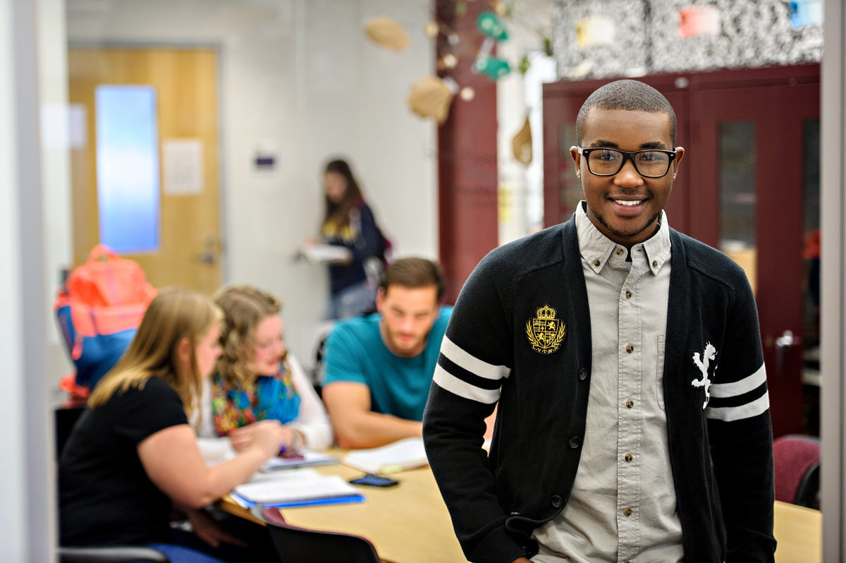 A confident college student looks at the camera while his classmates study behind him.