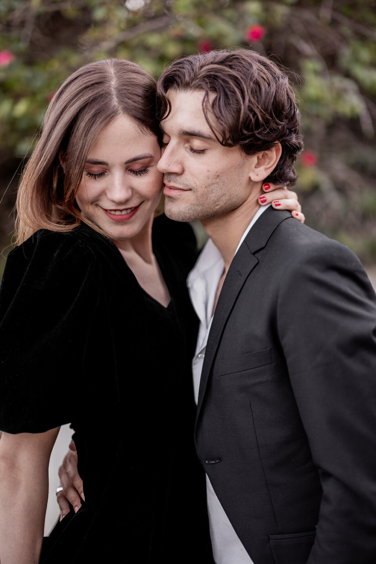 Couple embracing outdoors in soft natural light, sharing an intimate moment near flowering greenery.