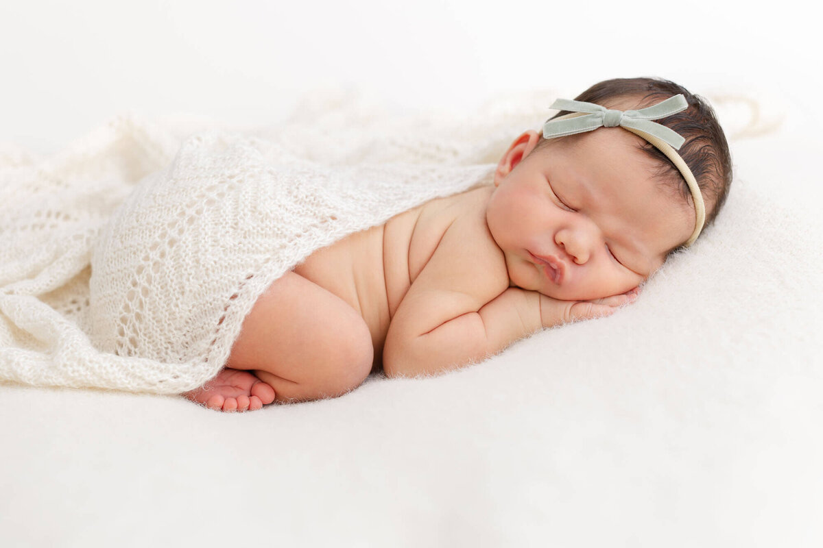 Newborn baby girl sleeping on belly with toes showing and one hand up under her head. She has a knit beige blanket draped over her back and is wearing a green velvet bow on head.