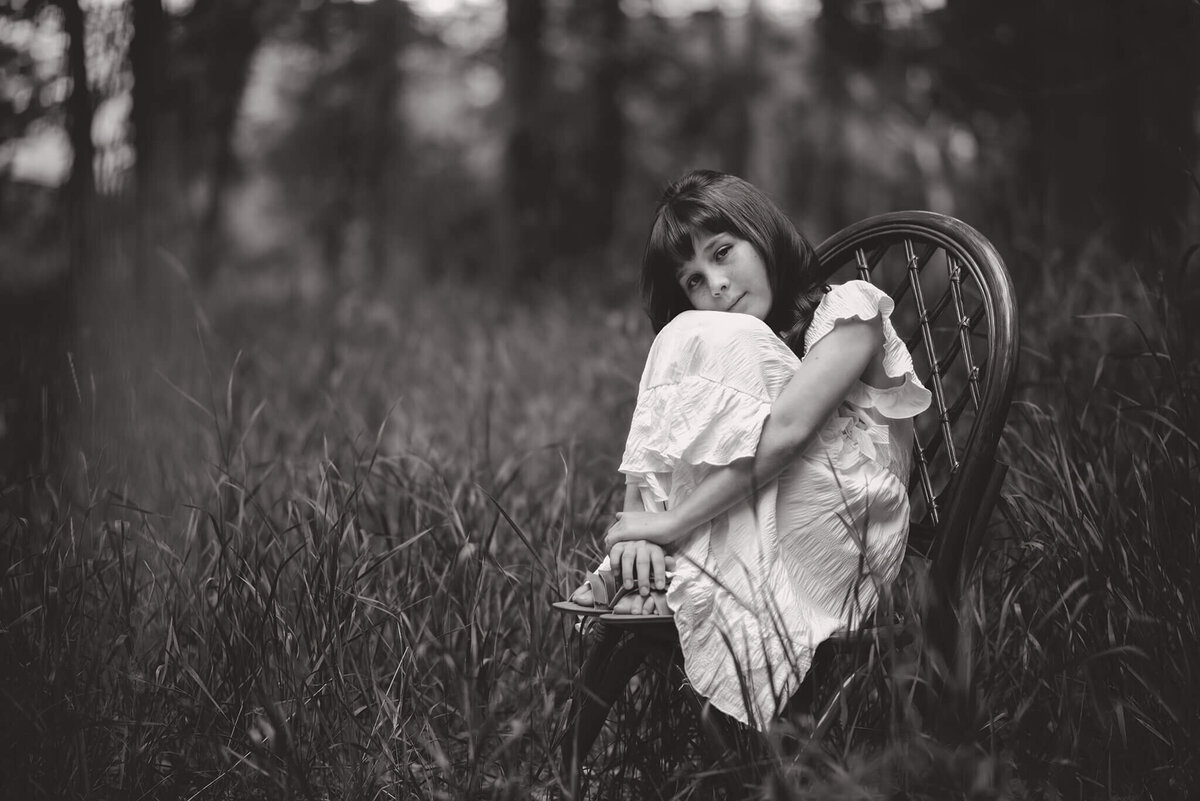 A little girl in a white dress clutches her knees while sitting on a wooden chair in the tall grass.  Black and white portrait by Brittany Danielle